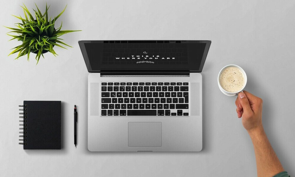 open laptop, with a cup of coffee, a book, a pen, and a plant on a table